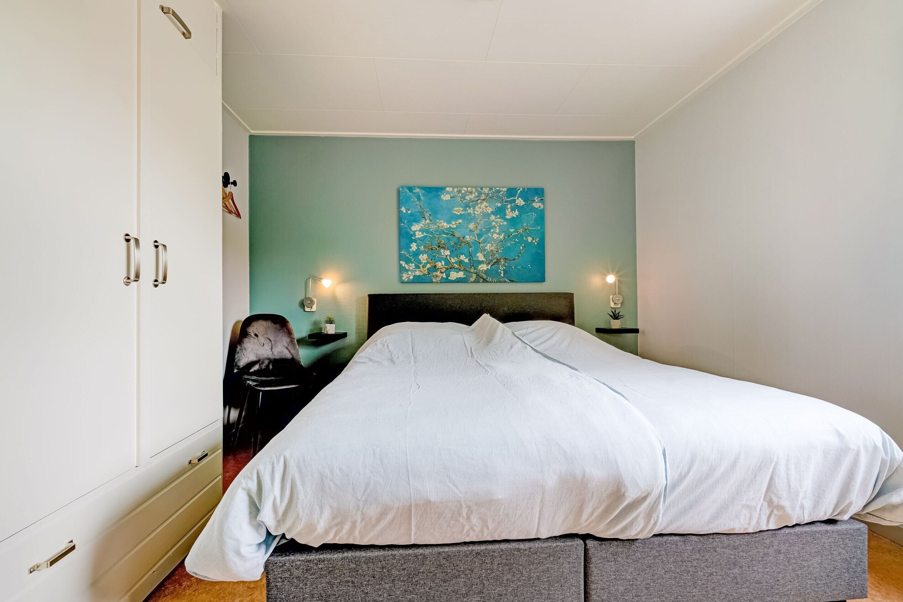 Een slaapkamer met een bed, nachtkastje, lamp en een schilderij aan de muur.