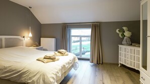 Une chambre avec un grand lit, une table de chevet, un radiateur, une fenêtre avec des rideaux, une armoire blanche et une vue sur l’extérieur.
