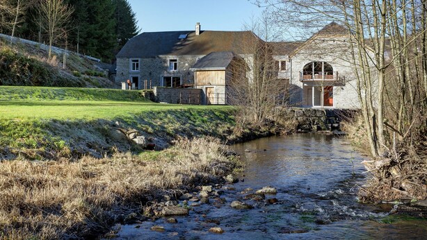 Une maison en pierre, avec une rivière devant, entourée d’arbres et d’une zone herbeuse.