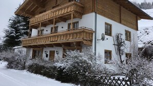 A two-story wooden chalet with a balcony, surrounded by snow-covered trees and bushes.