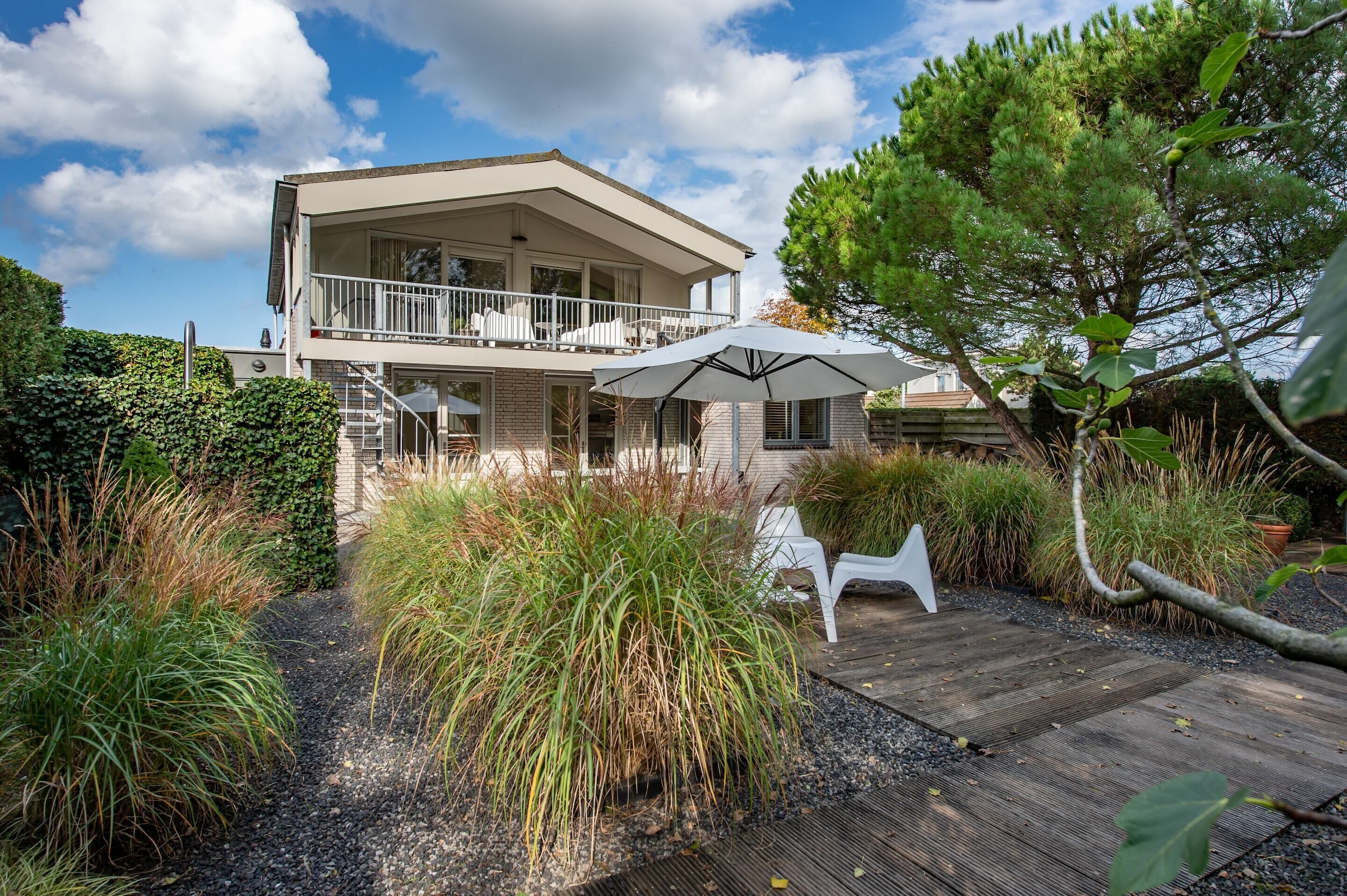 A two-story house with a balcony, surrounded by a garden with various plants and a wooden deck.
