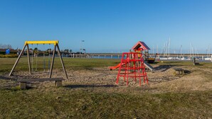 A playground with swings and a slide, a small red playhouse, and a sandy area with grass. - Bungalow Near Marina and Beach Kamperland (Kamperland)