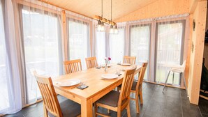 Ein Essbereich mit Holztisch und Stühlen, großen Fenstern mit leichten Vorhängen und einer Hängeleuchte.