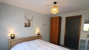 A bedroom with a wooden headboard, a large bed with white bedding, a wall-mounted deer painting, a wooden wardrobe, and a bathroom with a mirror and sink. - Holiday Home in Ardennes With Sauna & Nature (Manhay)