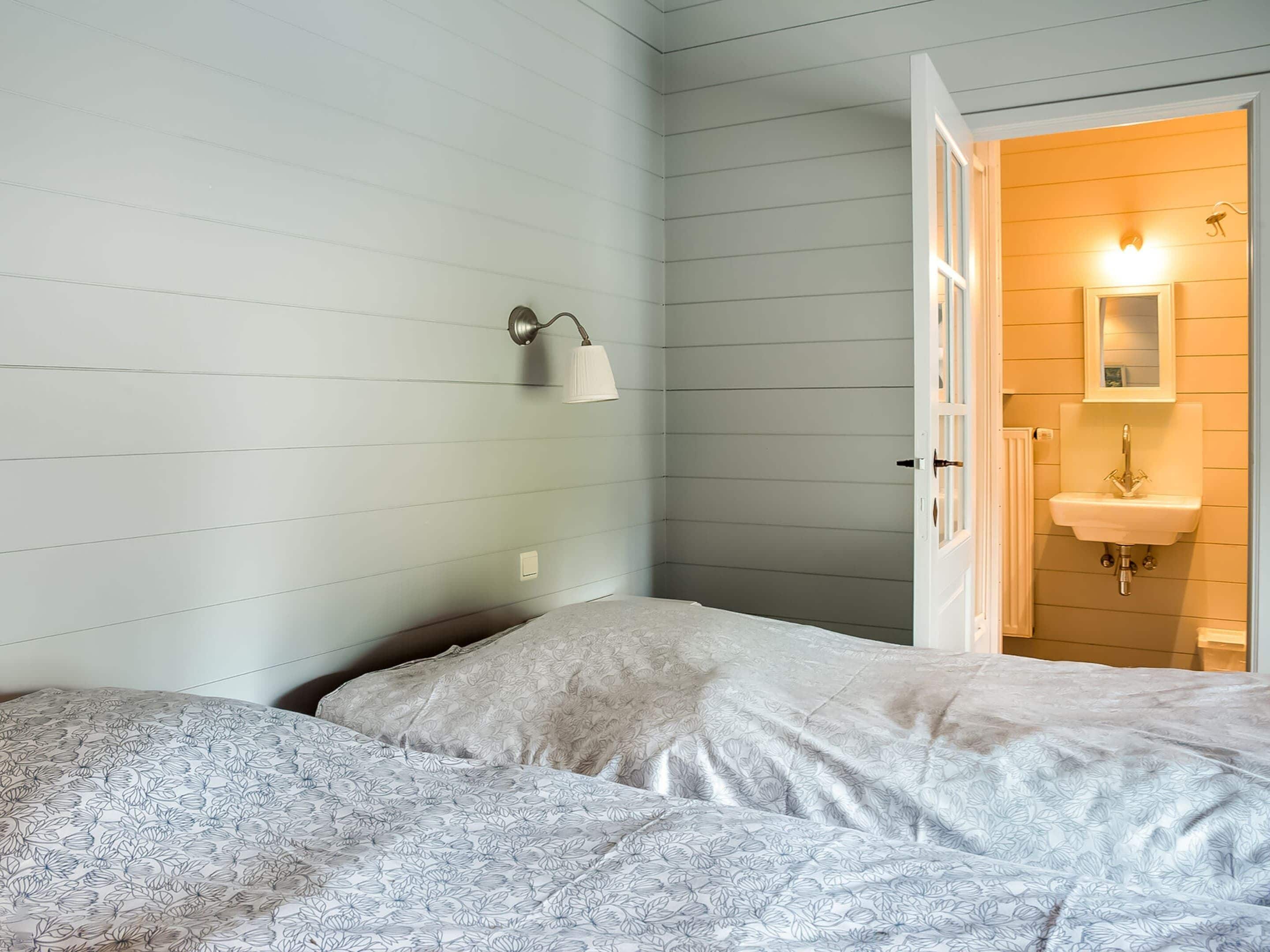 A bedroom with a bed, a nightstand, a wall-mounted lamp, and a bathroom visible through an open door.