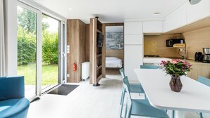 A modern hotel room with a large sliding glass door to a garden, a dining area with a white table and blue chairs, and a kitchenette with a coffee machine. - Chalet in Baalse Hei With Modern Comforts (Turnhout)