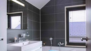A modern bathroom with dark tiles, a white vanity, a bathtub, and a window with blinds. - Nice Luxuriuos Villa With Sauna, Located in a Lake Area (Froidchapelle)