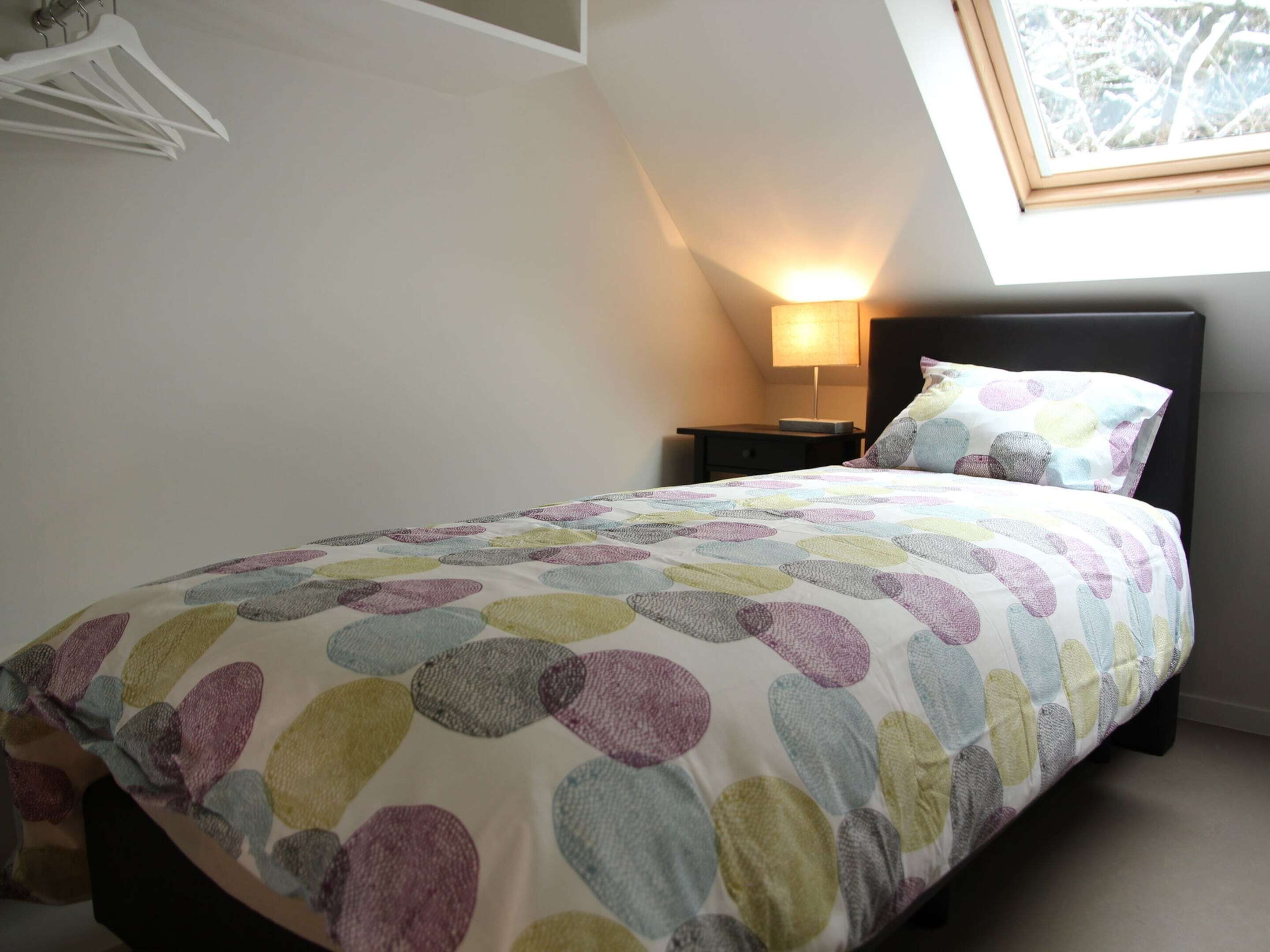 Un dormitorio con cama, mesita de noche, lámpara y ventana con vista a árboles.