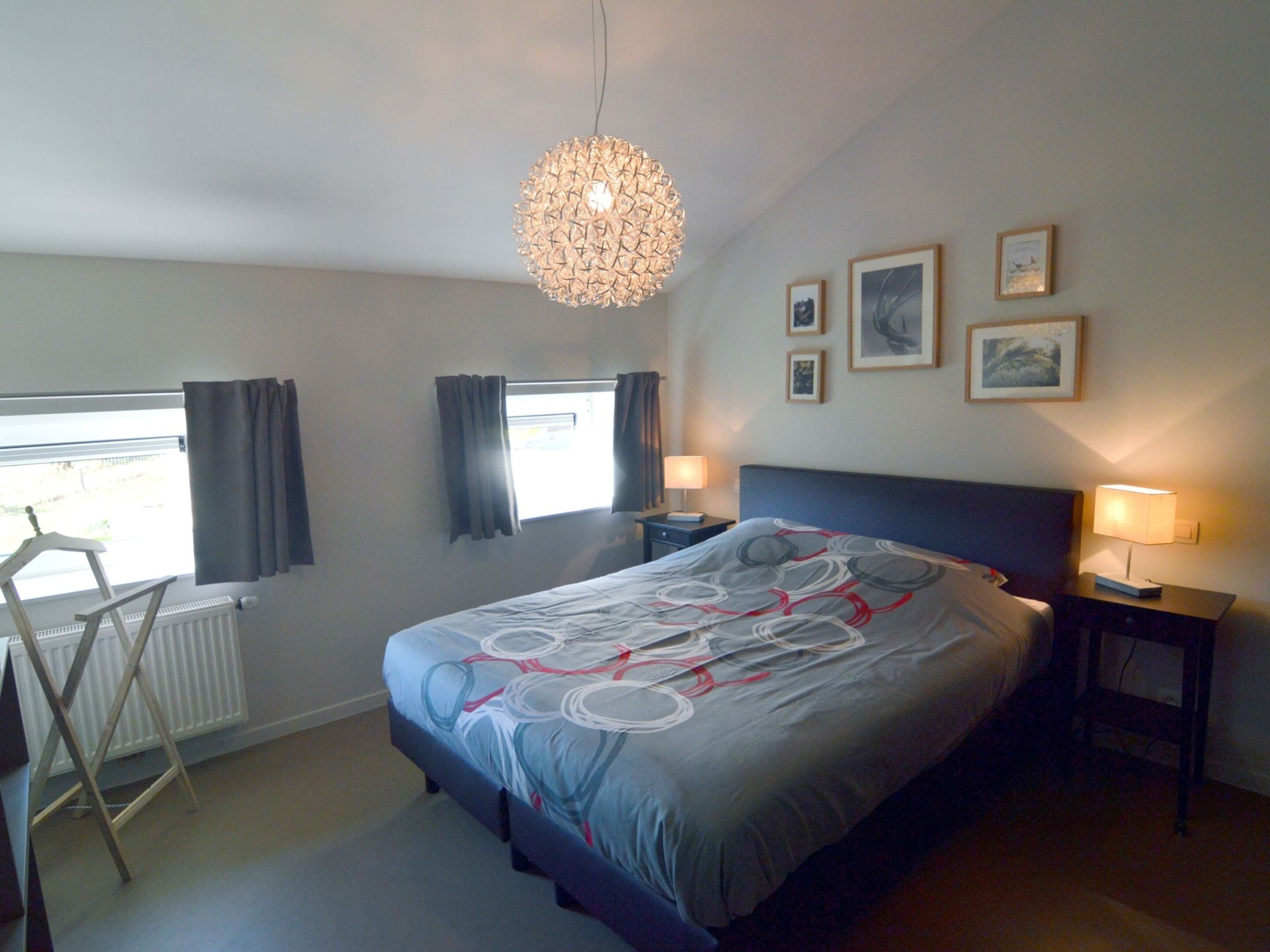 A bedroom with a bed, two bedside lamps, a radiator, a window with curtains, and a hanging light fixture.