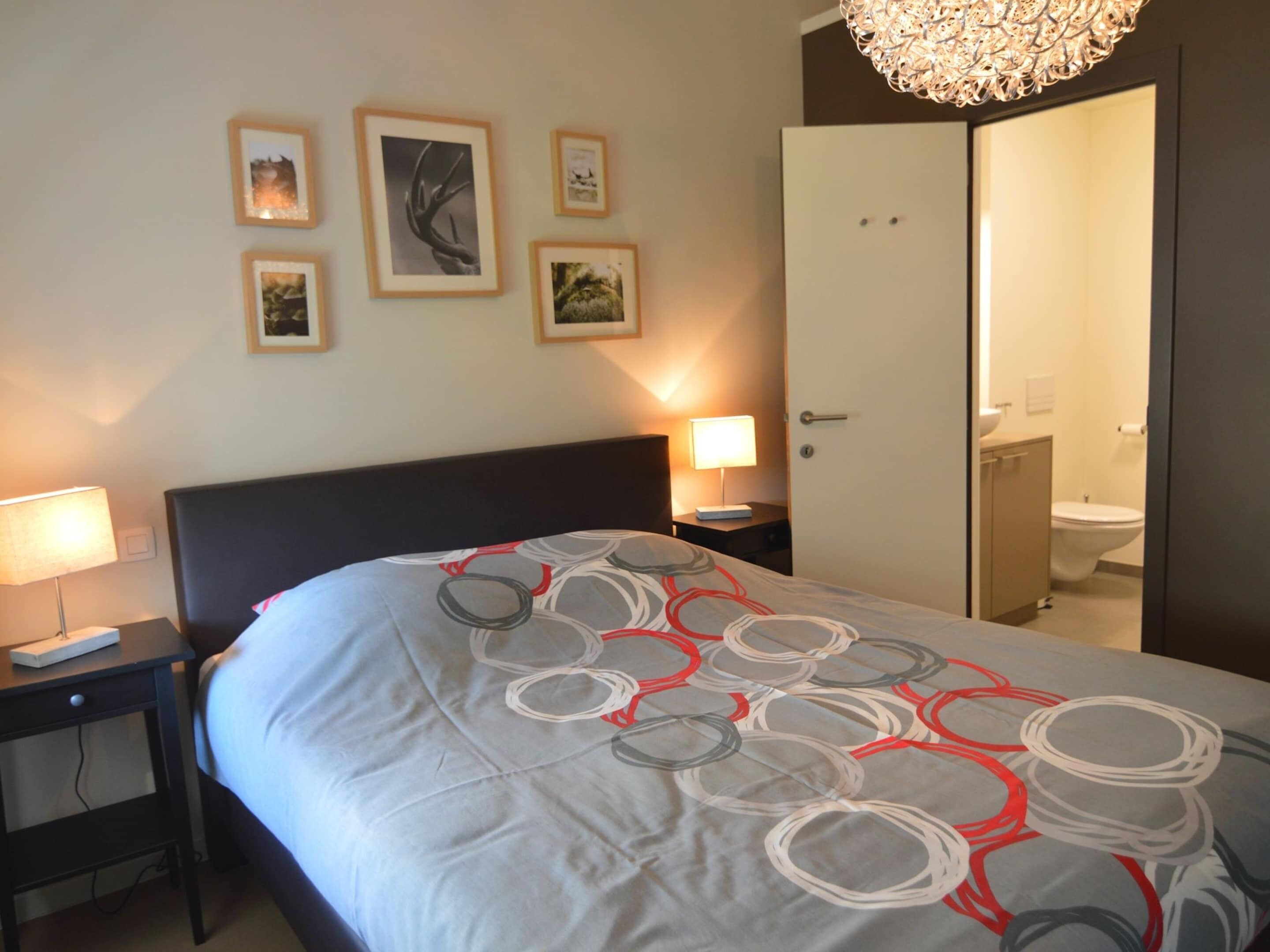 A hotel room with a bed, bedside tables, a lamp, a door to the bathroom, and wall-mounted pictures.