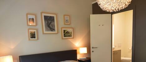 A hotel room with a bed, bedside tables, a lamp, a door to the bathroom, and wall-mounted pictures.