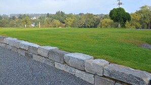 A stone wall bordering a grassy field with trees and houses in the background.