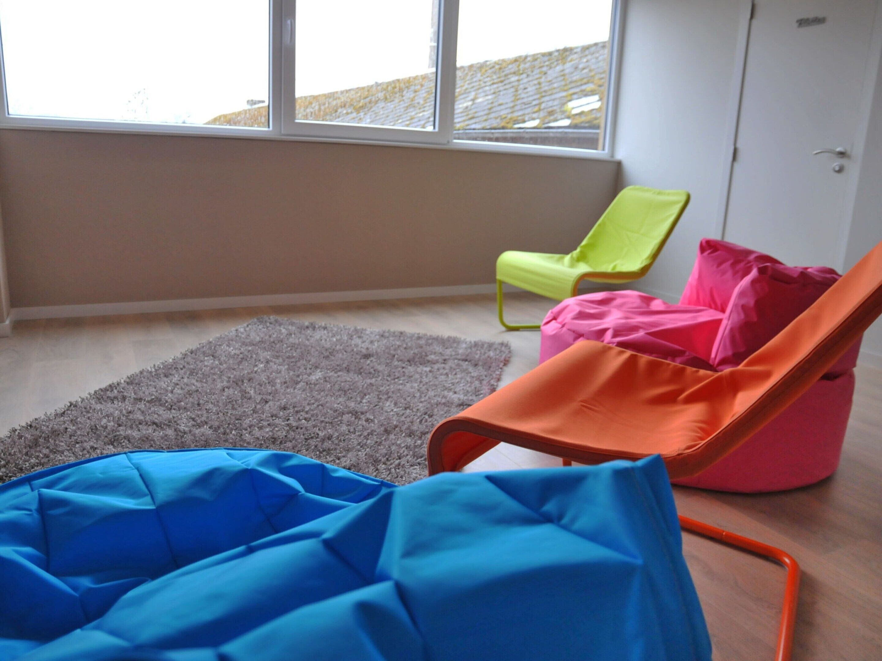 Une pièce avec une grande fenêtre, un tapis gris et des poufs colorés.