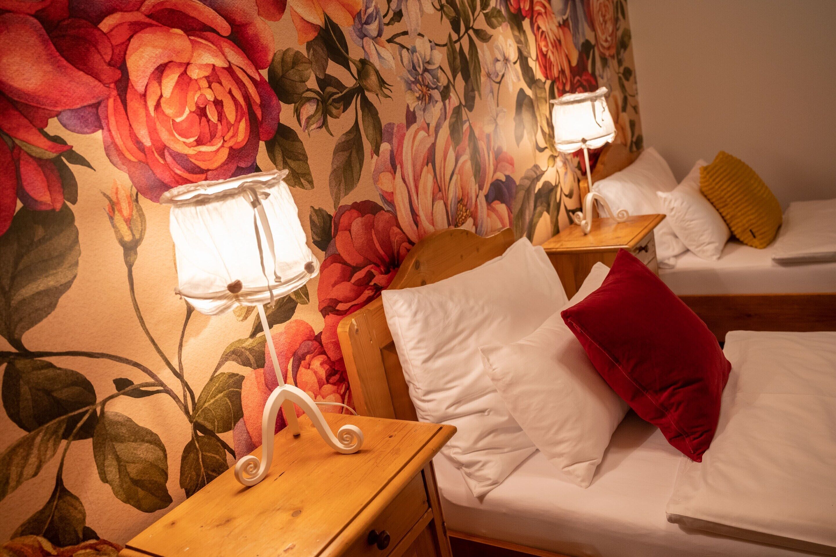 A hotel room with two beds, floral wallpaper, bedside lamps, white bedding, and a red pillow.