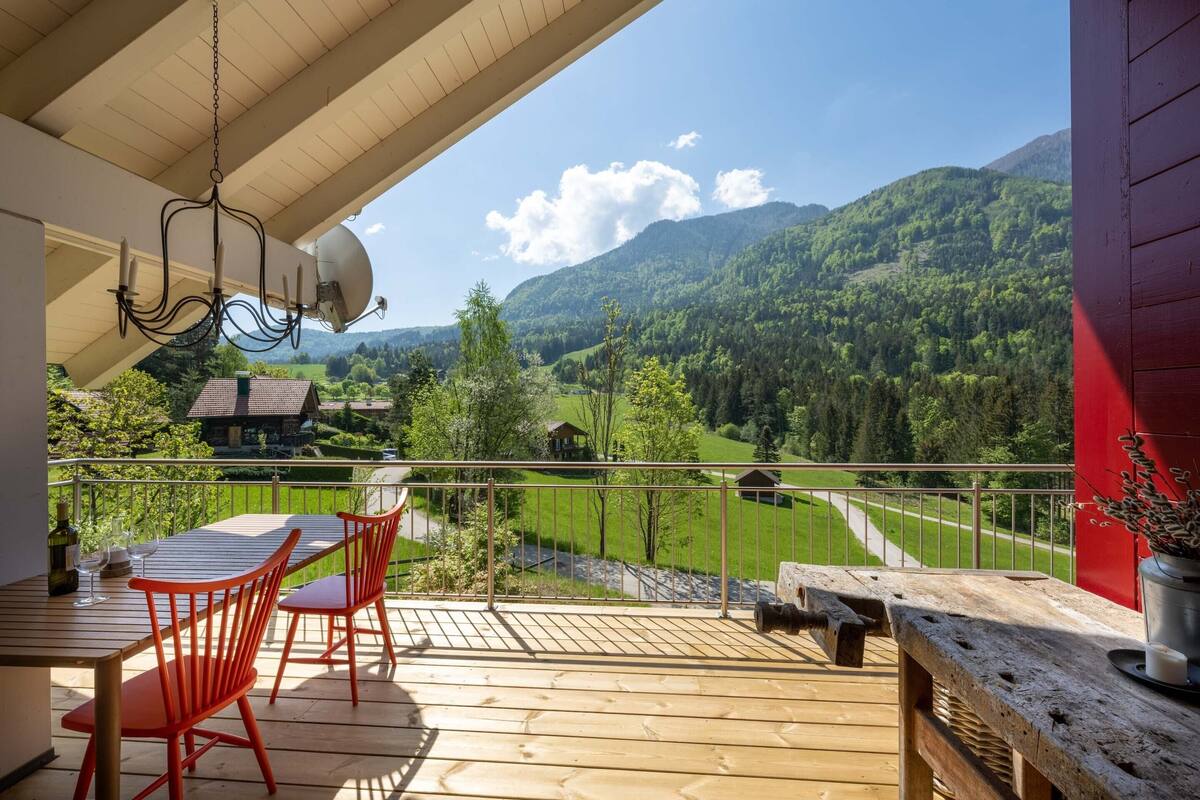 A wooden deck with a table and chairs, a hanging light fixture, and a view of a green valley and mountains.