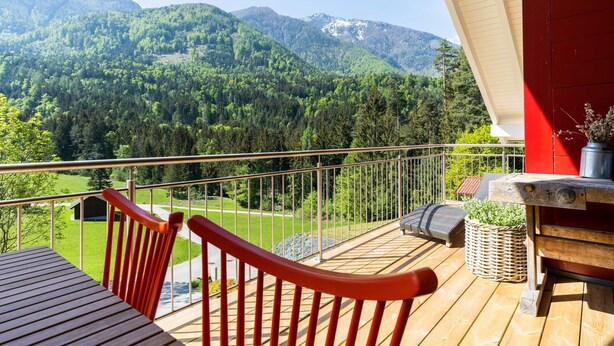 Um deck de madeira com guarda-corpo vermelho, com vista para uma paisagem montanhosa com florestas densas e céu claro.