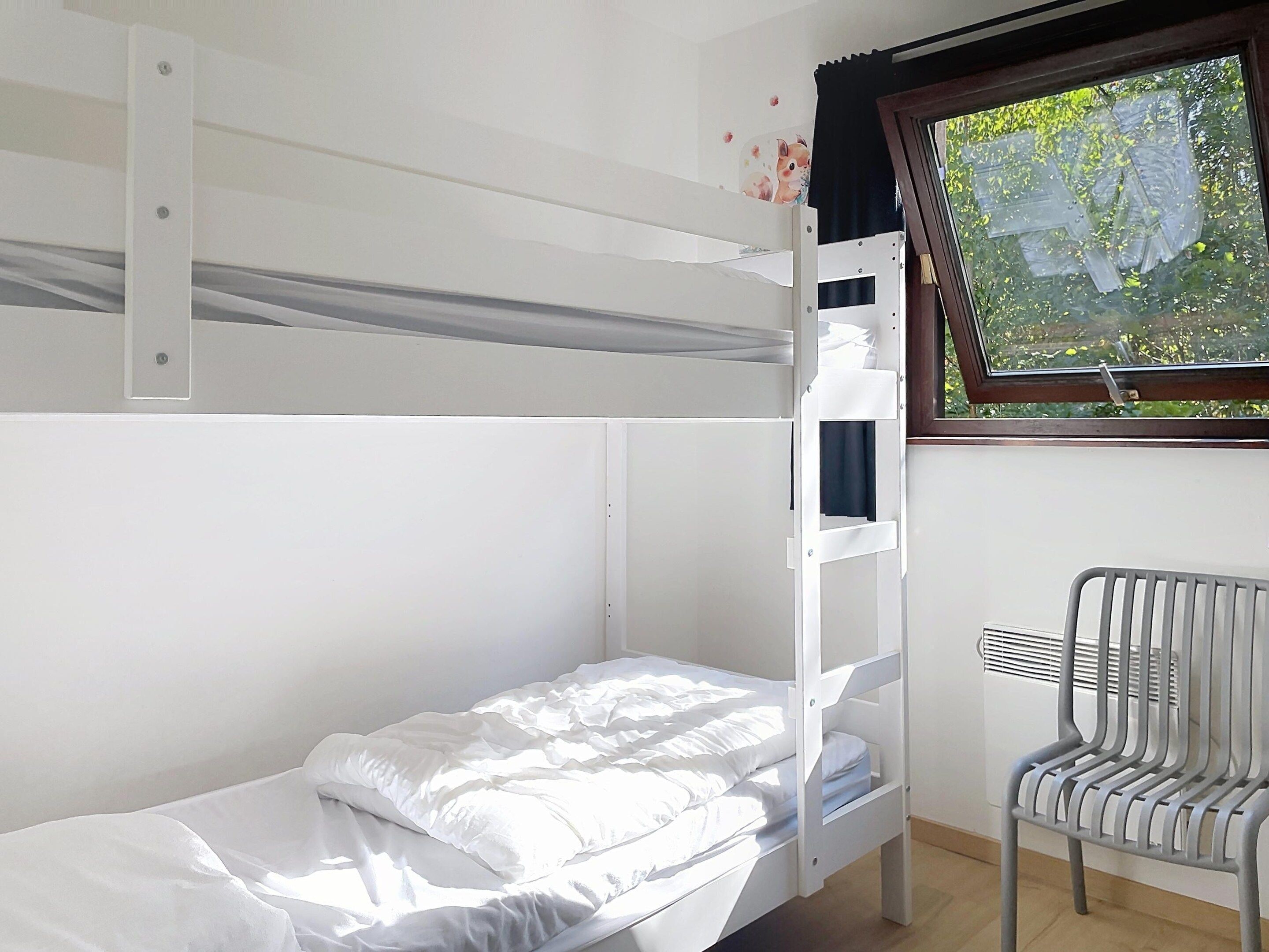 Uma cama de beliche com uma cama inferior e uma cadeira ao lado de uma janela com vista para o verde.