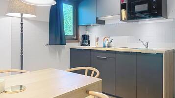 Cozinha compacta equipada com micro-ondas, chaleira e máquina de café. Inclui uma mesa de jantar com cadeiras e um abajur.