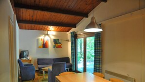 A cozy room with a wooden ceiling, a dining table, chairs, a sofa, a television, and a window with curtains. - Chalet in Belgium Surrounded by Nature (Viroinval)
