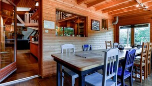 Interior de uma cabana de madeira, com área de jantar, cozinha e escada para um andar superior.
