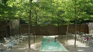 Una piscina esterna con due persone a nuotare, circondata da sedie e tavoli bianchi, con alberi e un muro di pietra sullo sfondo.
