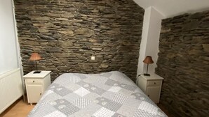 A bedroom with a stone wall, a bed with a patterned bedspread, two bedside tables with lamps, and a radiator. - Holiday Home in Semois Valley Near Kayaking (Vresse-sur-Semois)