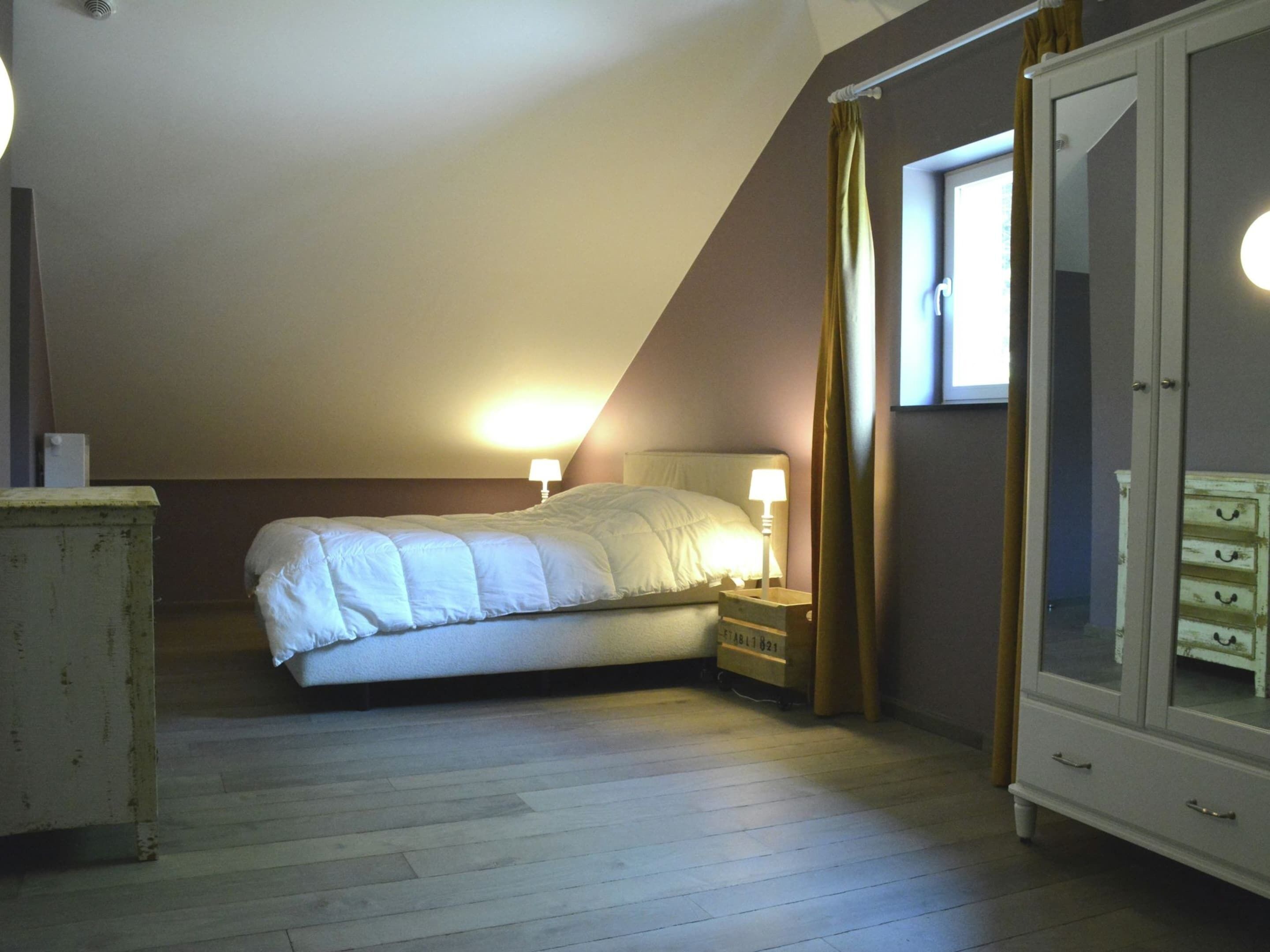 Een slaapkamer met een bed, bedlampje, dressoir en een raam met gordijnen.