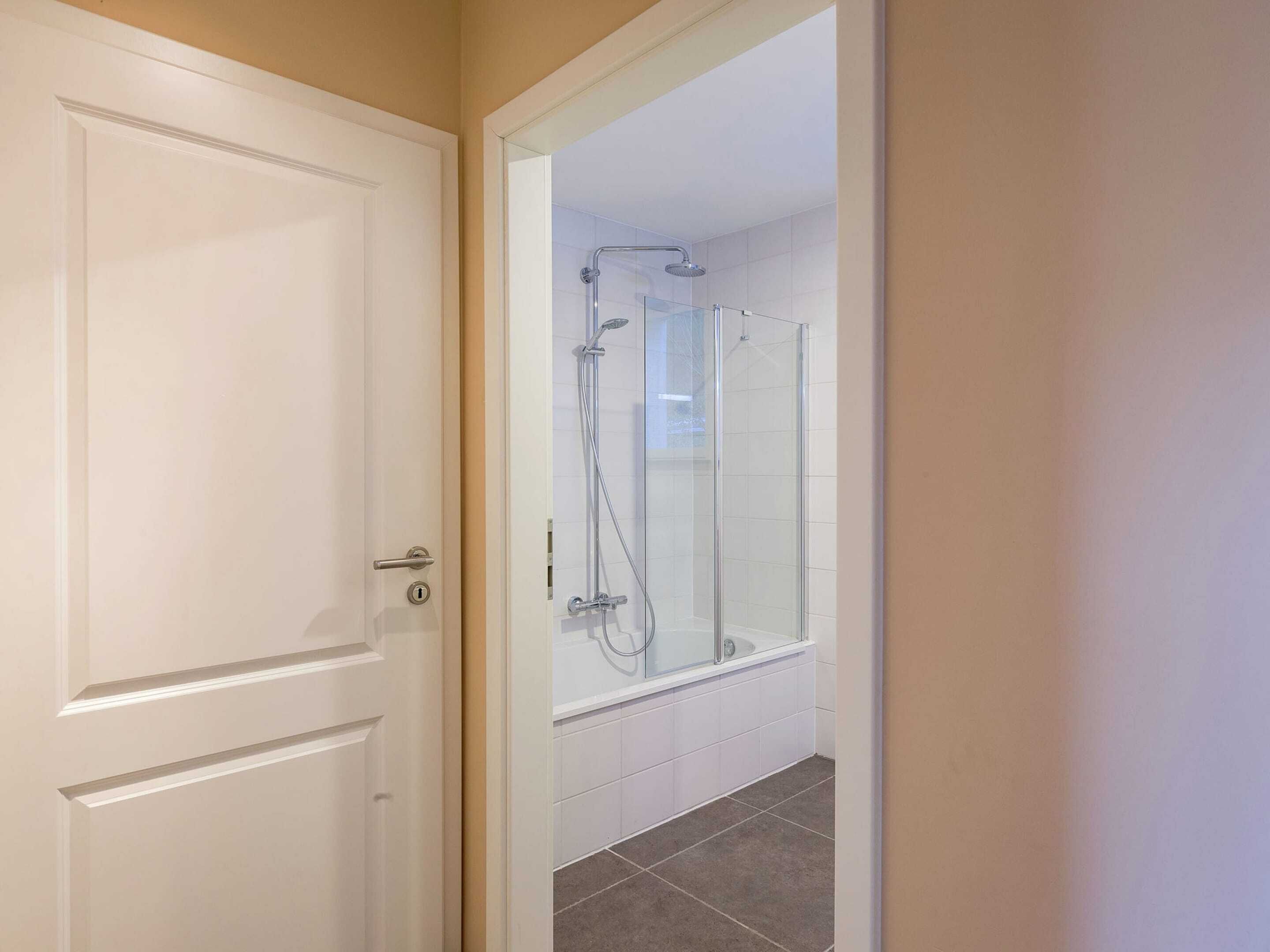 A bathroom with a glass shower enclosure, white tiles, and a door with a handle.