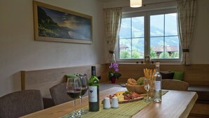 Ein Essbereich mit einem Holztisch, der für eine Mahlzeit gedeckt ist, mit einer Weinflasche, Gläsern, einer Käseplatte und Blick ins Freie durch ein Fenster.