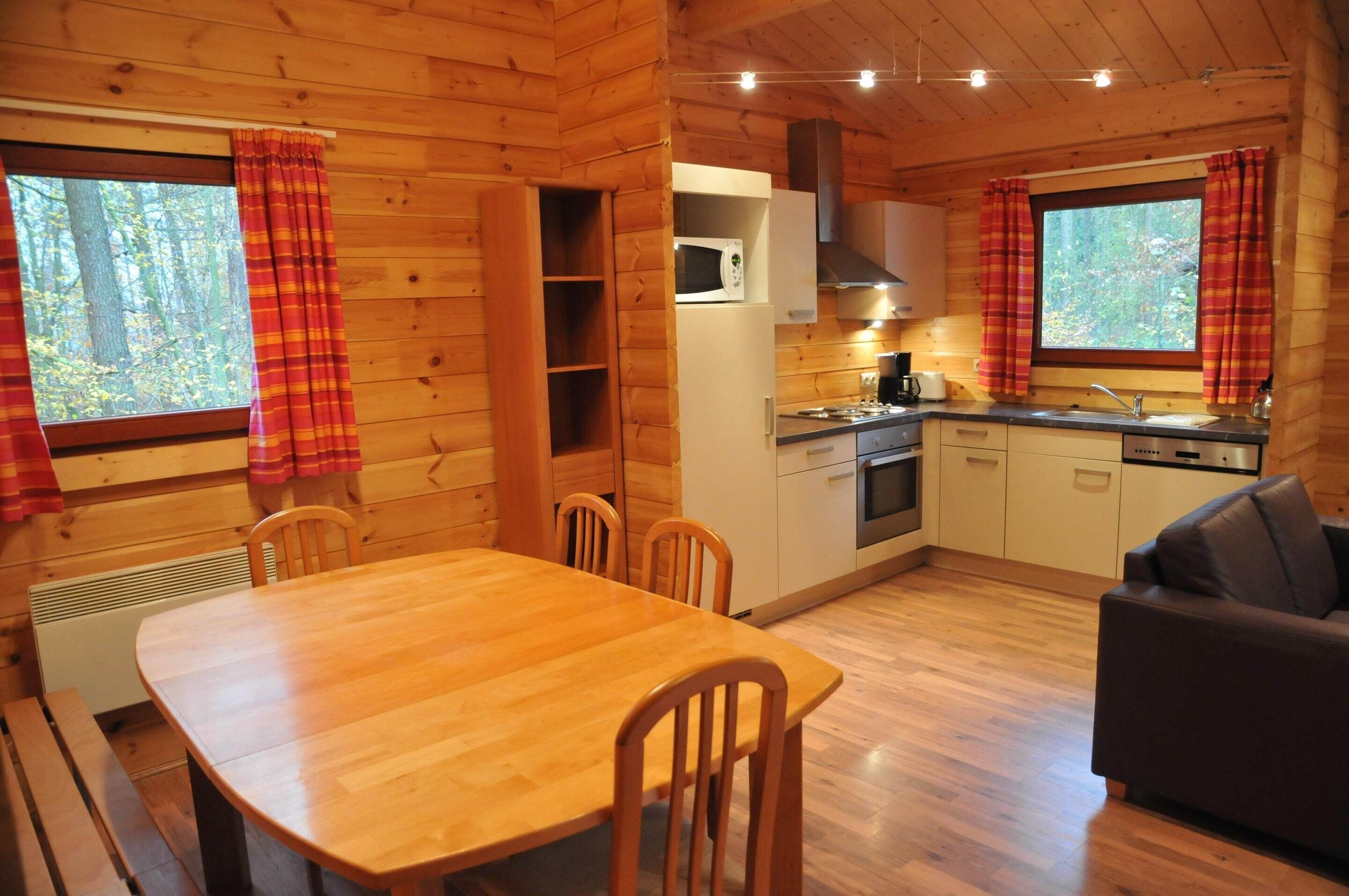 A wooden cabin interior with a dining table, chairs, a sofa, a kitchen with a microwave, stove, and cabinets.