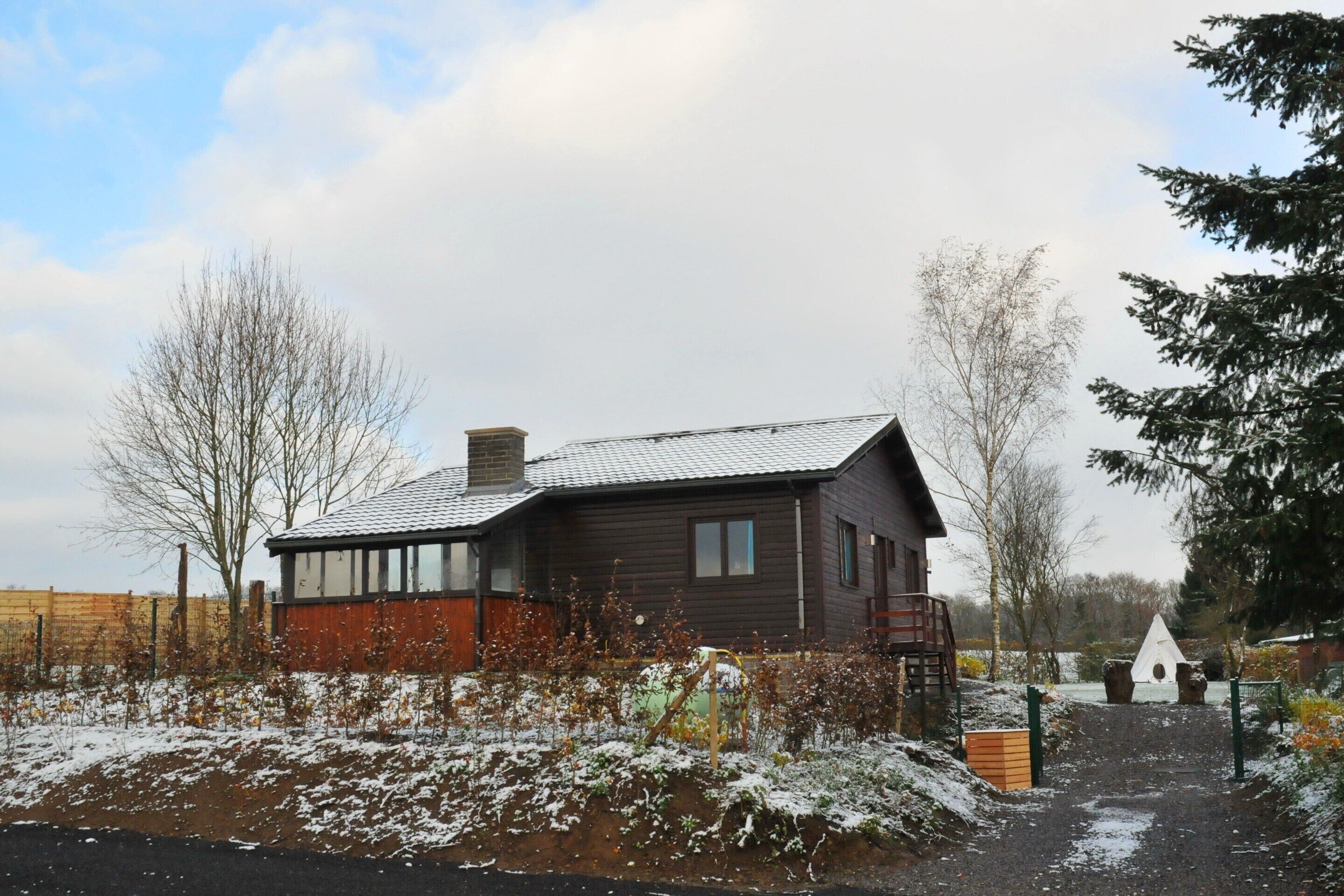 A wooden house with a sloped roof, a visible chimney, and a fenced garden area with snow on the ground.