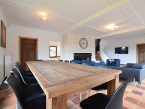 A spacious living area with a large wooden table, blue sofas, a fireplace, a clock, and a TV mounted on the wall.