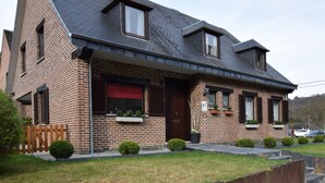 A two-story brick house with a dark roof, a front porch with a bench, and a small front yard with a wooden fence and potted plants. - Cozy Family Holiday Home (La Roche-en-Ardenne)