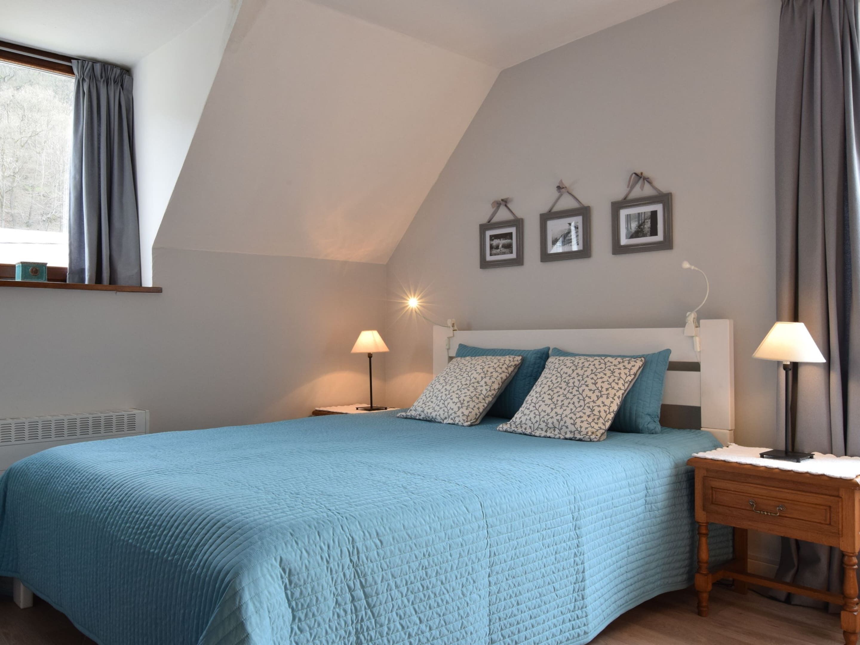 A bedroom with a bed, bedside lamps, a nightstand, a radiator, and a window with curtains.