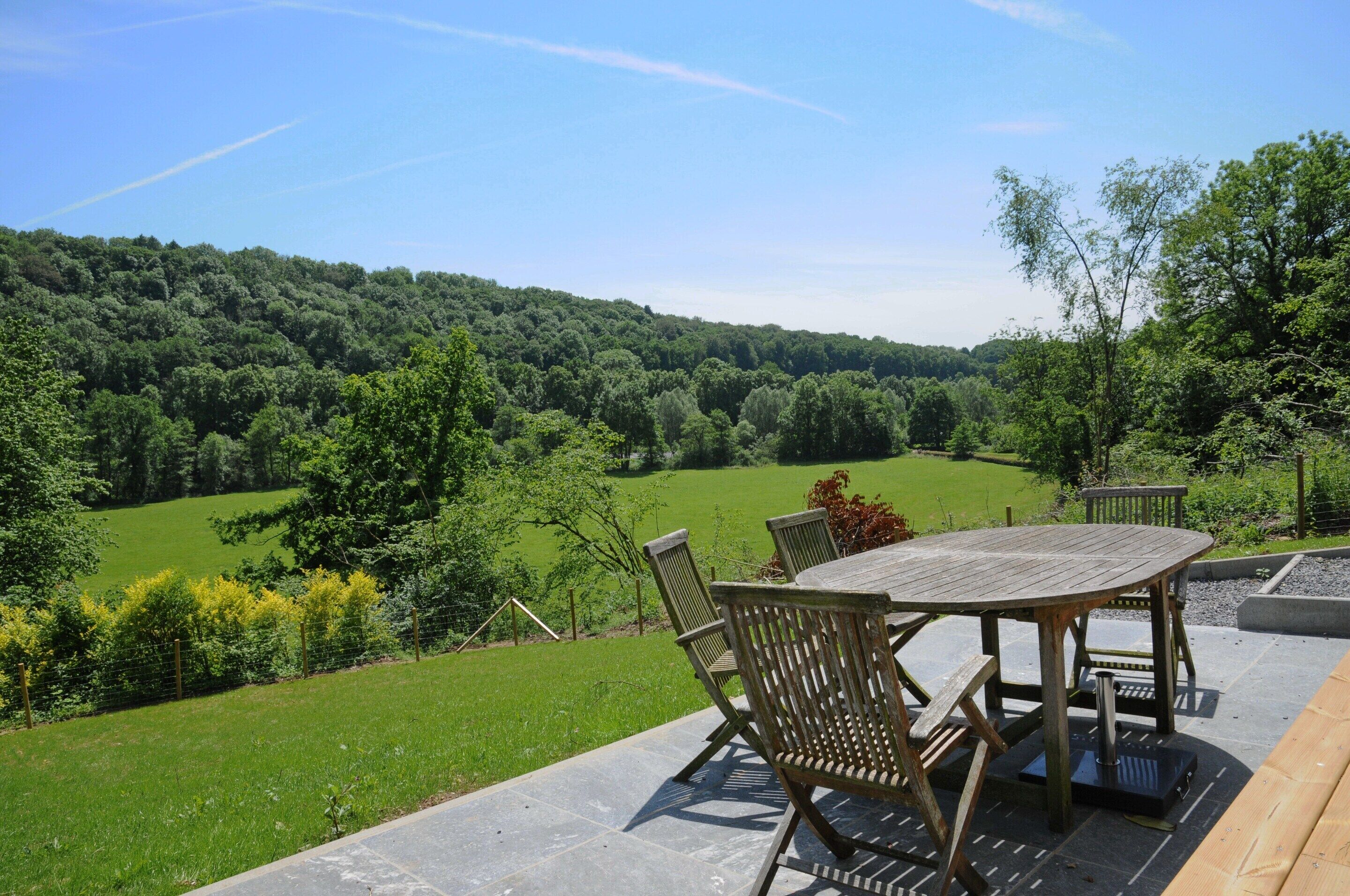 Une terrasse extérieure avec une table et des chaises en bois, entourée d’un paysage verdoyant et luxuriant, et avec une colline boisée en arrière-plan.
