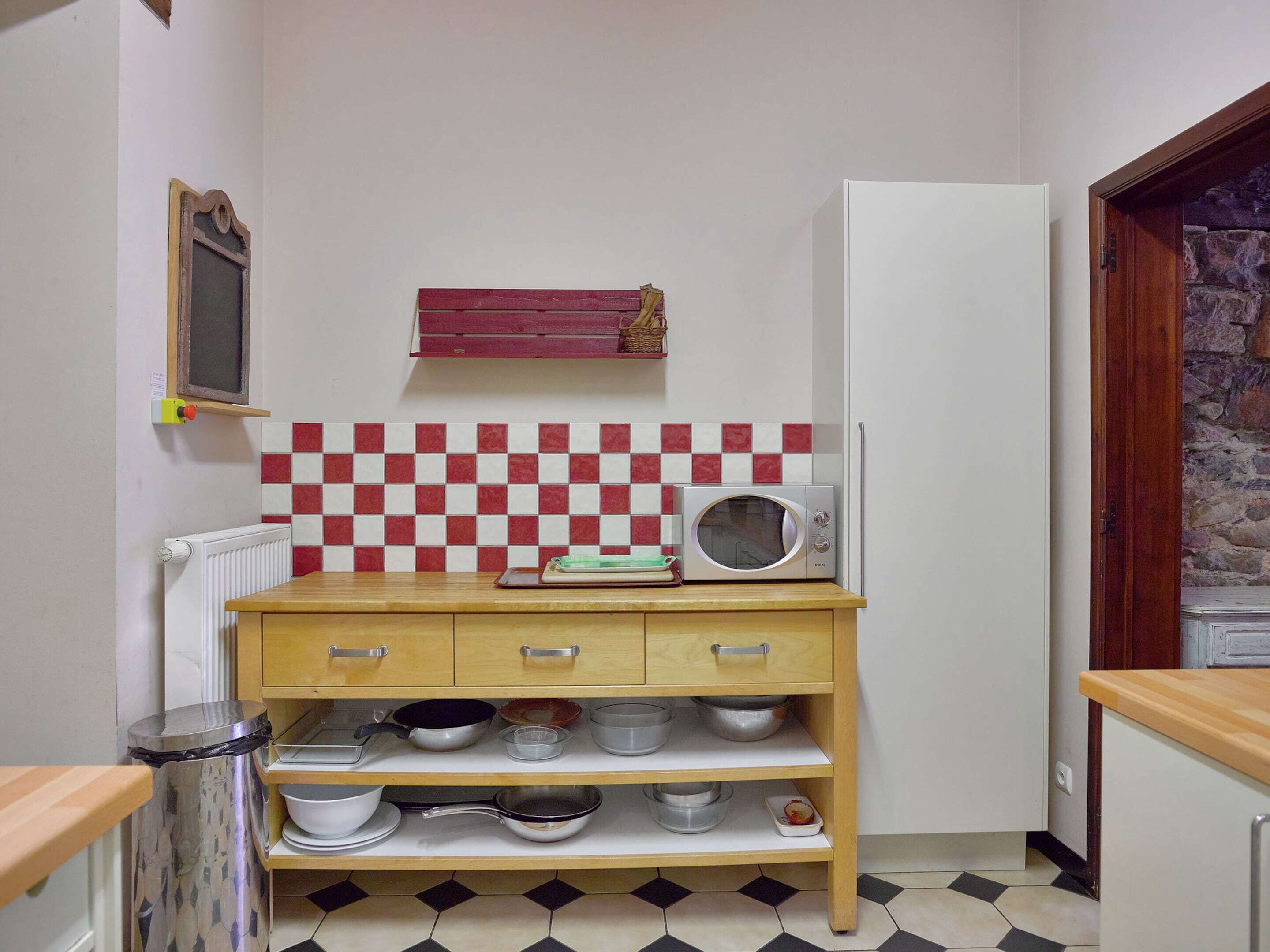 A compact kitchen with a microwave, shelves with cookware, a red and white checkered backsplash, and a stone wall door.