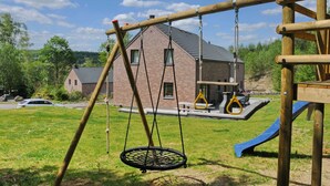 A playground with a swing set, a slide, and a brick building in the background. - Holiday Home in Barvaux With Sauna & Garden (Durbuy)