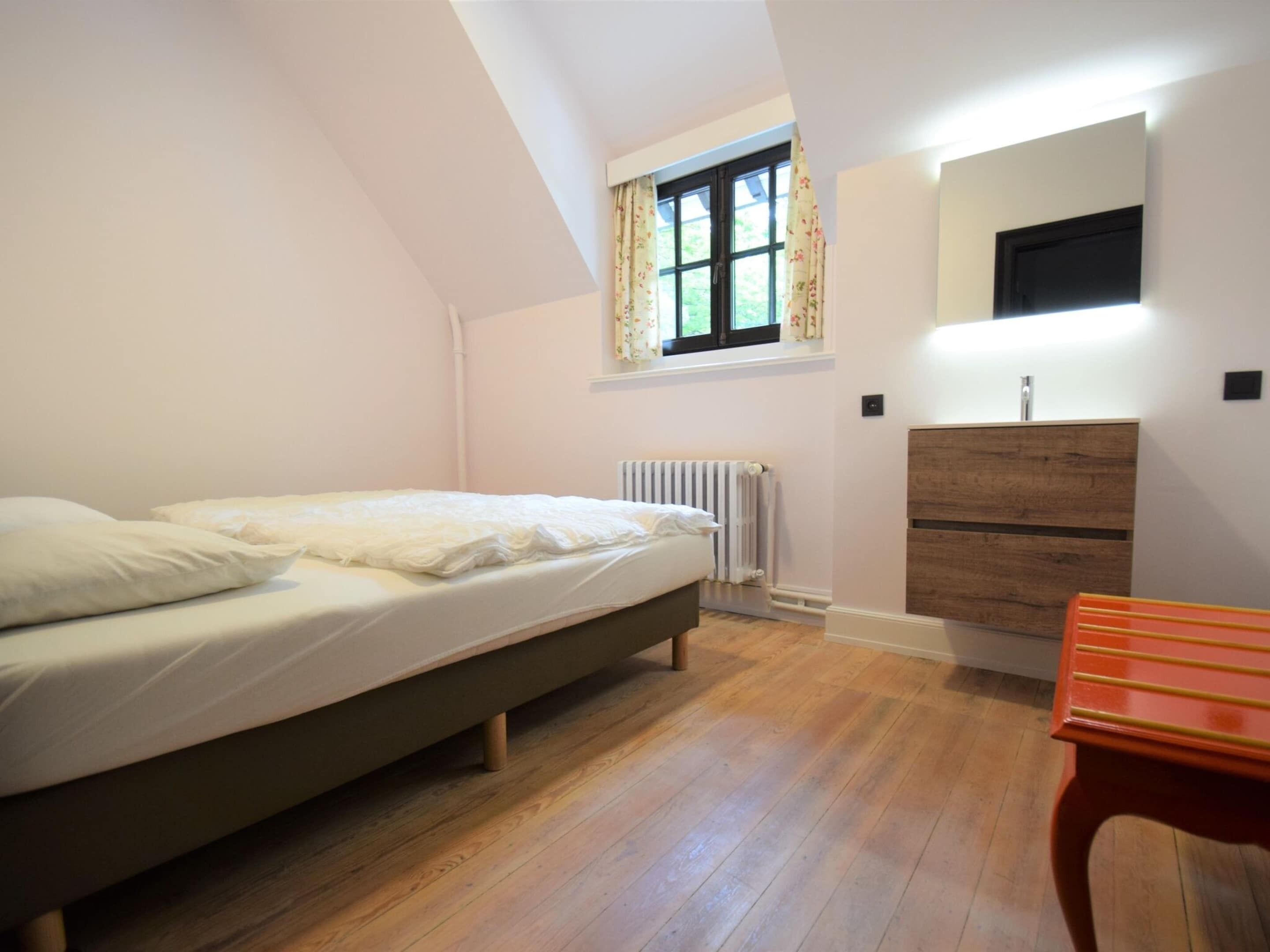 A bedroom with a bed, a wooden dresser, a mirror, and a window with curtains.