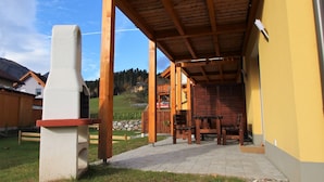 Un área de estar al aire libre techada, con mobiliario de madera, un pilar de piedra y vistas a una montaña y casas al fondo.