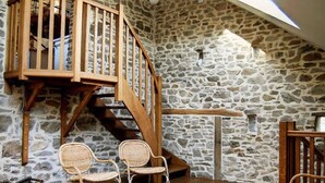 Una habitación rústica con pared de piedra, escalera de madera, sillas de mimbre y una mesa de madera.