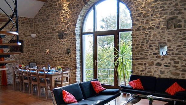 Una habitación amplia con pared de piedra, un gran ventanal arqueado, un comedor y un sofá de cuero negro con cojines rojos.