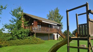 Une structure de jeu en bois avec un toboggan, une terrasse en bois et un bâtiment doté d’un balcon et d’un escalier, le tout sur fond de paysage.