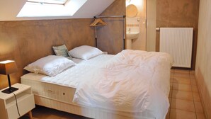 Une chambre avec un puits de lumière, un lit avec une literie blanche, une table de chevet avec une lampe, un porte-vêtements suspendu et un radiateur.