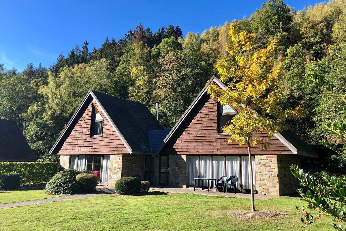 A two-story lodge with a stone facade, a covered patio with outdoor seating, and a forested backdrop.