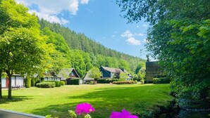 A serene lakeside view with a wooden deck, vibrant pink flowers, and a row of houses with sloped roofs. - Cottage Famille - Family Resort With Wellness in the Heart of the Ardennes (Malmedy)