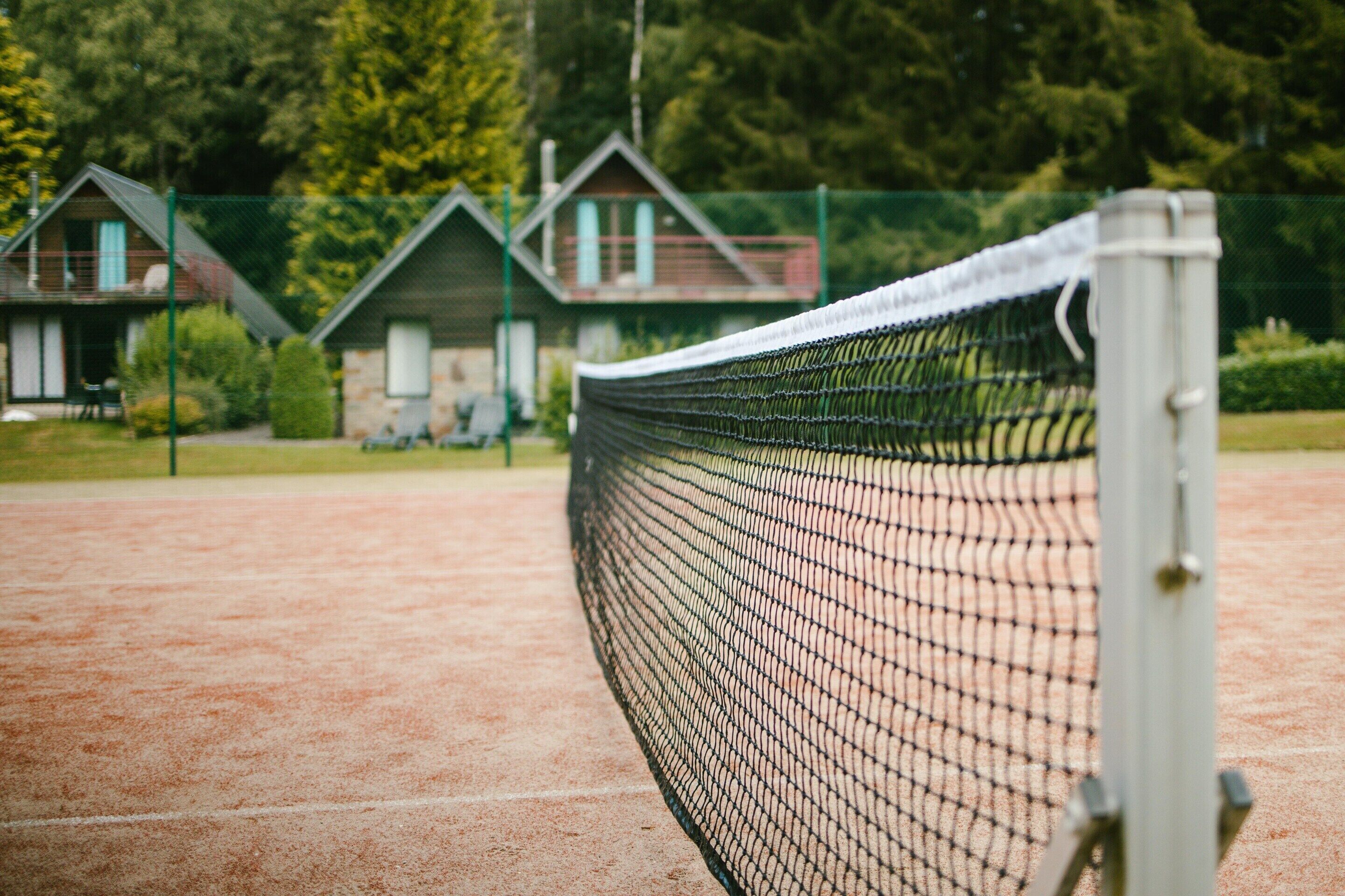 A tennis court with a net, surrounded by a fence and a few small buildings in the background.