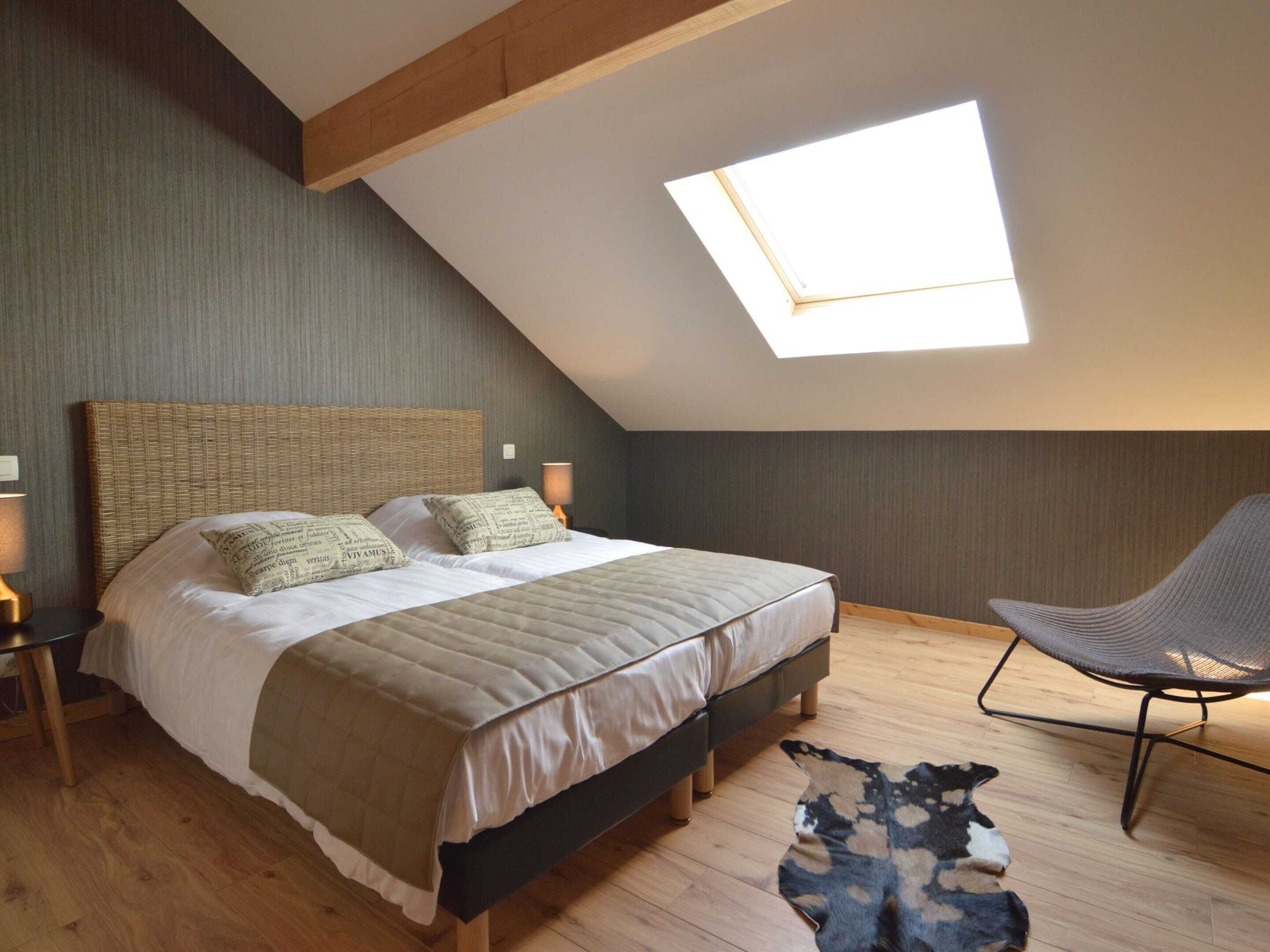 Een slaapkamer met een schuine kap, een groot bed met een geweven hoofdbord, twee bedlampen, een stoel en een vloerkleed.