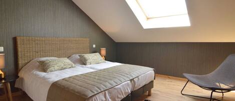 Een slaapkamer met een schuine kap, een groot bed met een geweven hoofdbord, twee bedlampen, een stoel en een vloerkleed.