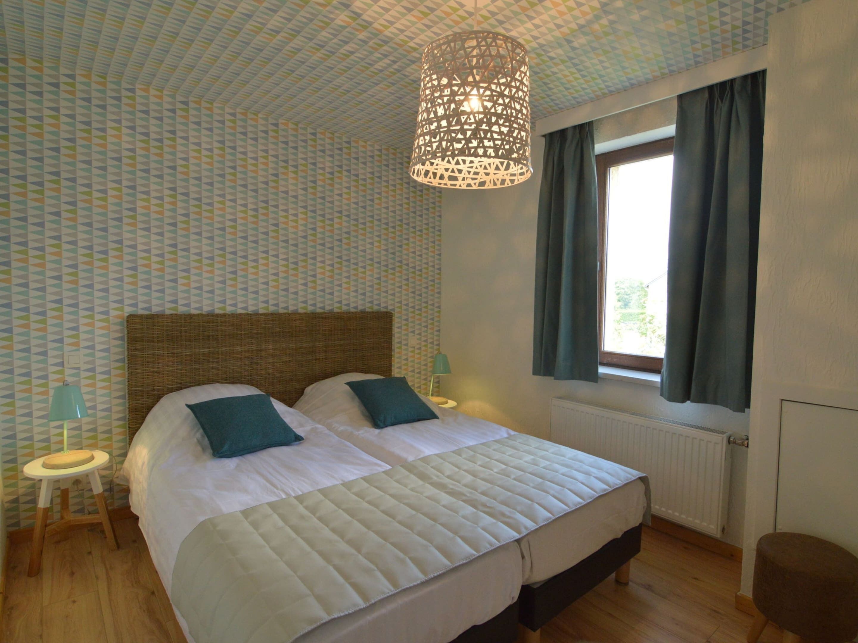 A bedroom with a large bed, two bedside tables, a hanging light fixture, a window with curtains, and a patterned wallpaper.