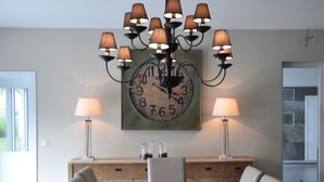 Uma área de refeições com uma mesa comprida, várias cadeiras, um candelabro, duas luminárias de parede e um relógio grande.